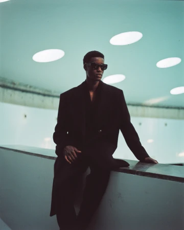Stylish African-American male model in black coat and sunglasses poses indoors against a pale aqua ceiling with round recessed lights.