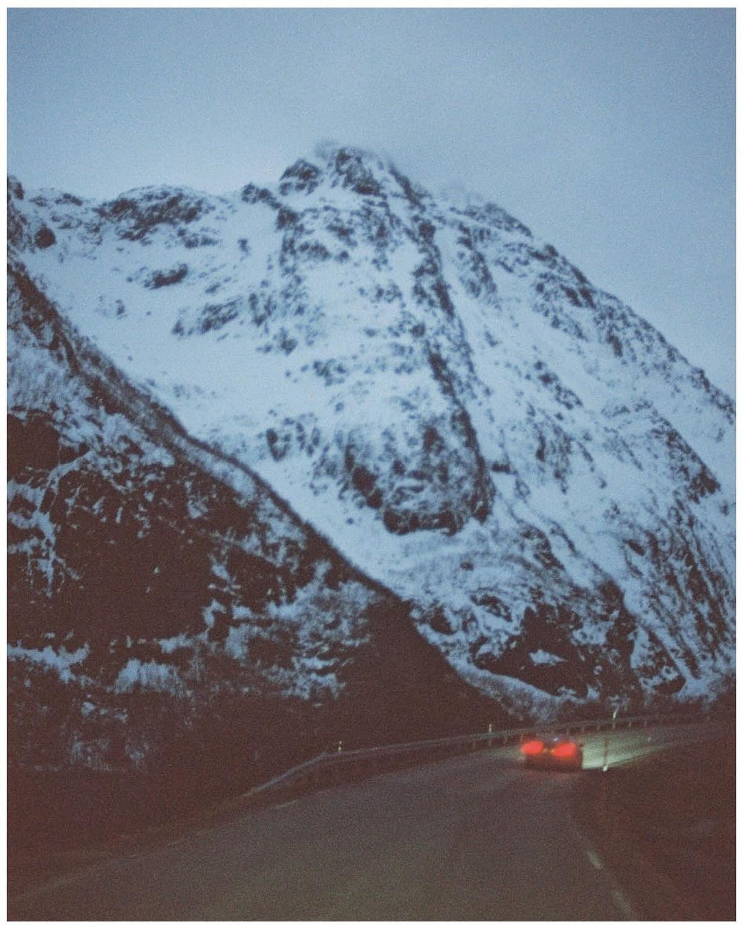 Snow-covered mountain dominates the landscape with a road leading to the base, illuminated by car headlights under an overcast sky.