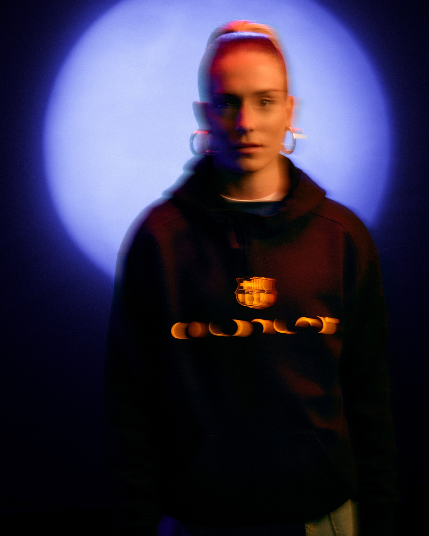 Portrait of a young adult female wearing a black FC Barcelona hoodie, posing in a dark studio with a blue spotlight behind her.