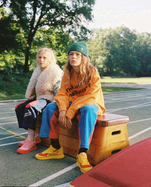 Two young children are sitting on a wooden bench in an outdoor setting, wearing colorful streetwear and sneakers in a lifestyle shot.