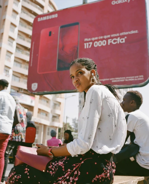 Young woman sits outdoors with a magenta purse against an ad for Samsung mobile phones in a bright, sunny, urban environment.