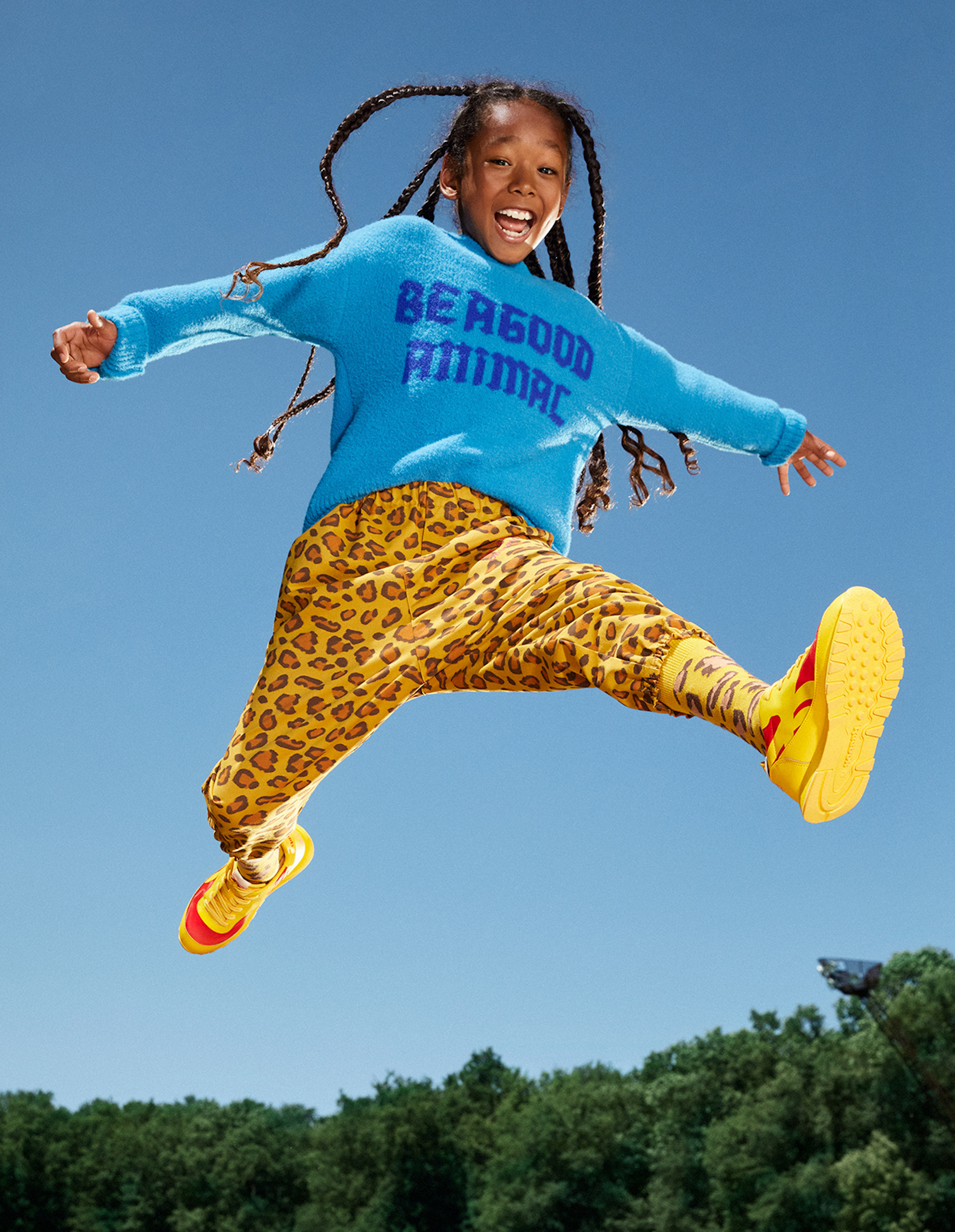 Girl with braids is mid-air jumping against a blue sky; wearing a blue sweater with text, leopard print pants, and yellow sneakers.