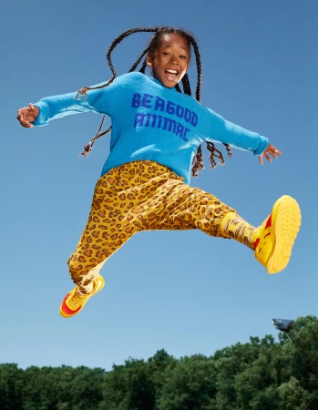 Girl with braids is mid-air jumping against a blue sky; wearing a blue sweater with text, leopard print pants, and yellow sneakers. 