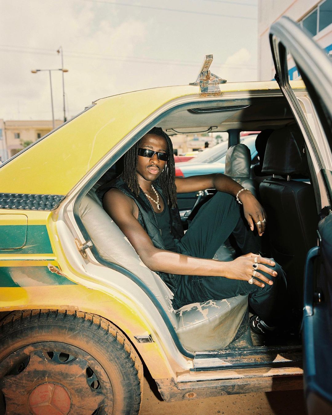 Adult male with sunglasses sits in the back of a yellow taxi wearing black tank top, pants, and jewelry in an outdoor, urban setting.