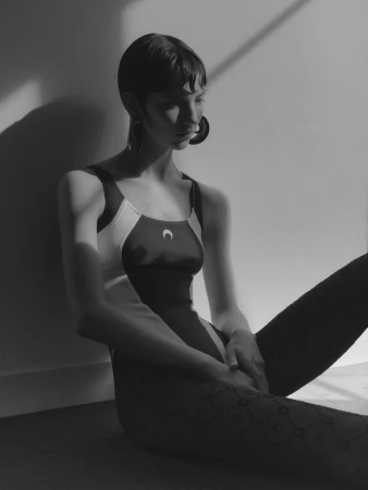 Model with short dark hair wearing a patterned leotard and large earrings seated against a plain wall in sharp lighting.