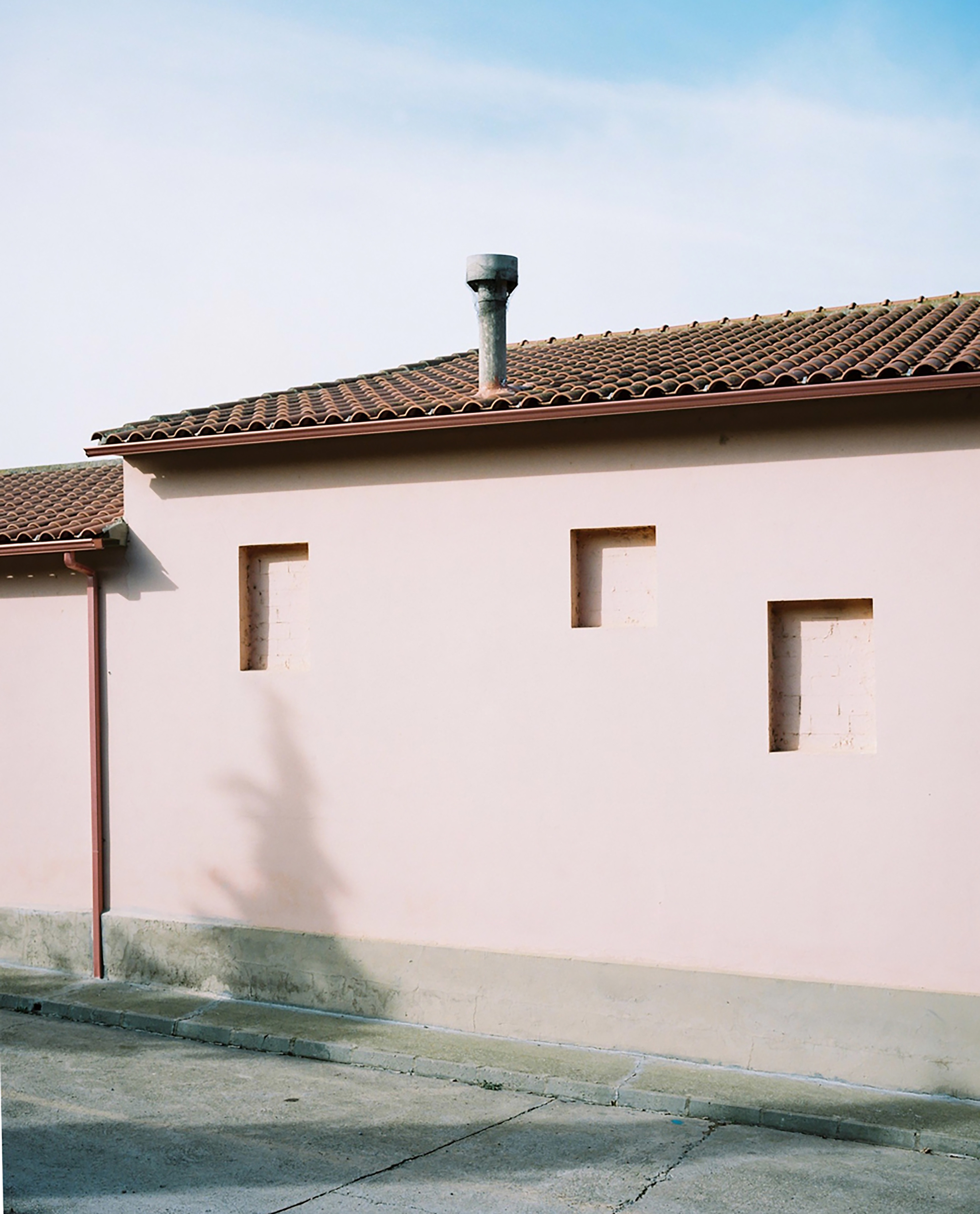 Exterior of a pale pink building with a tiled roof, chimney, and rectangular recessed openings under a blue sky. 