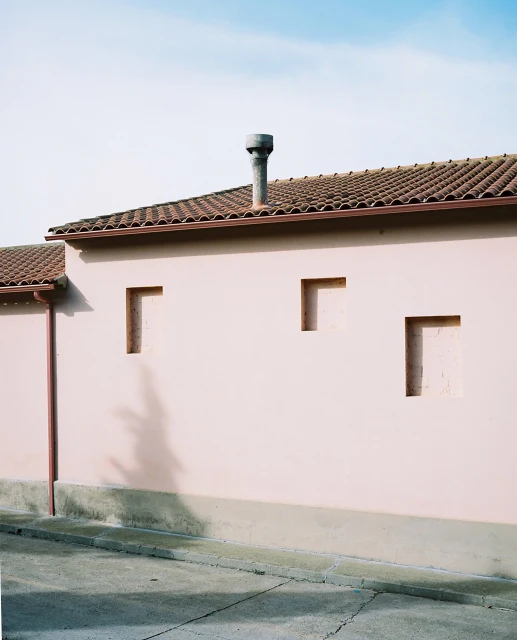 Exterior of a pale pink building with a tiled roof, chimney, and rectangular recessed openings under a blue sky. 