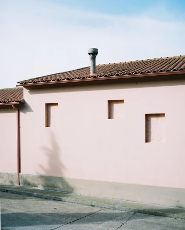 Exterior of a pale pink building with a tiled roof, chimney, and rectangular recessed openings under a blue sky. 
