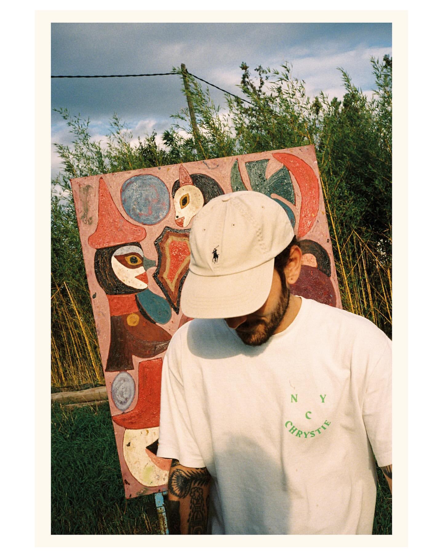 Young man in cap and white tee stands outdoors in front of colorful folk art painting, green foliage, and sky with clouds.