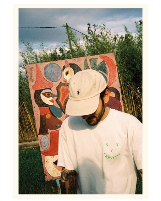 Young man in cap and white tee stands outdoors in front of colorful folk art painting, green foliage, and sky with clouds.