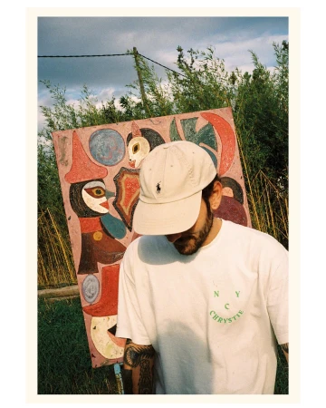 Young man in cap and white tee stands outdoors in front of colorful folk art painting, green foliage, and sky with clouds.