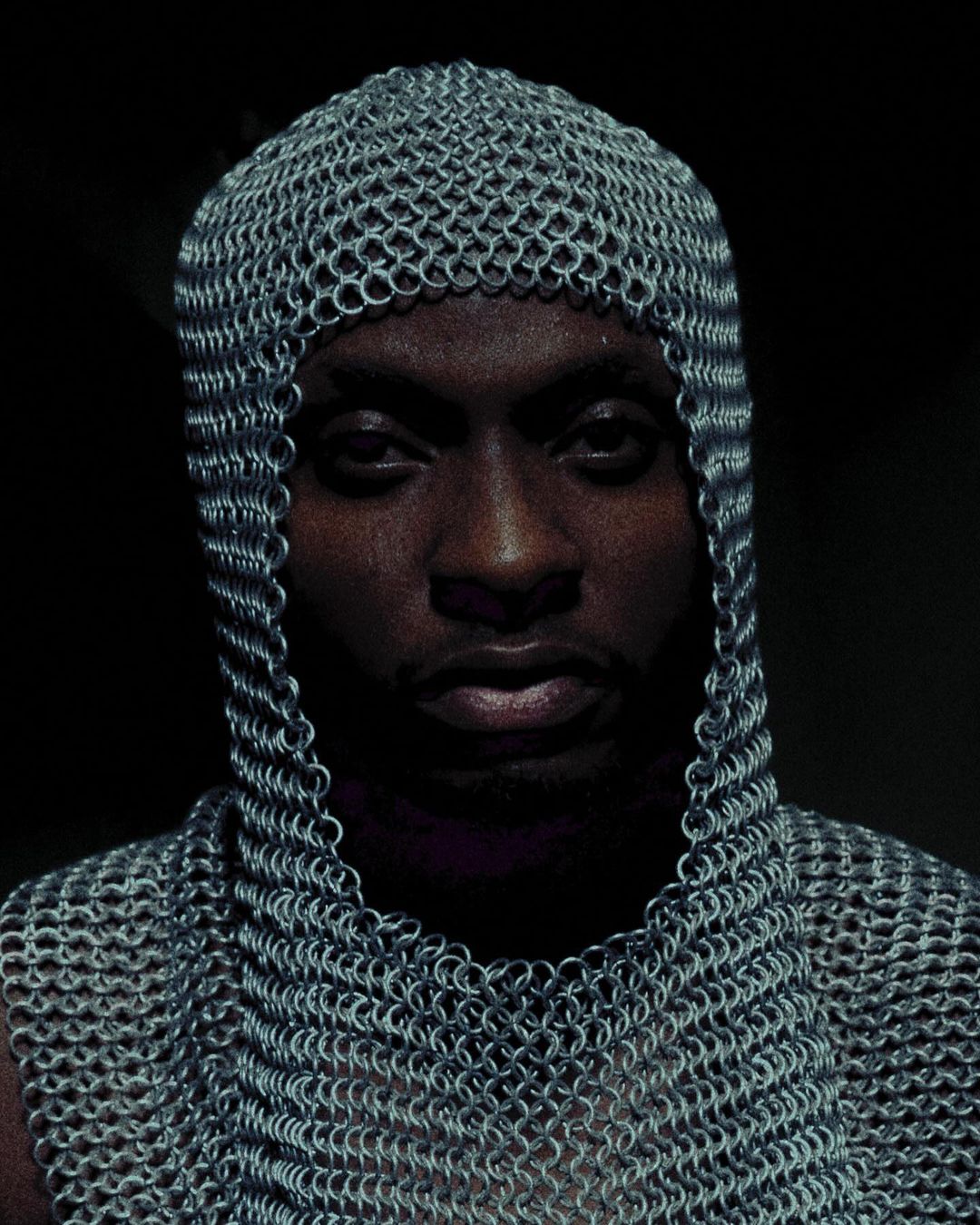 Portrait of an adult male wearing a chainmail coif and shirt, looking directly at the camera against a dark background.