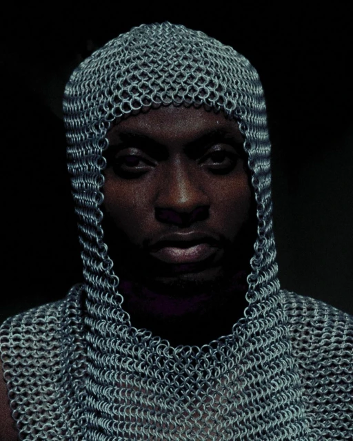 Portrait of an adult male wearing a chainmail coif and shirt, looking directly at the camera against a dark background.