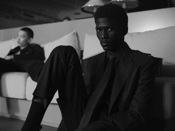 Black and white portrait of a black man wearing a dark suit seated on the floor with another man seated on a sofa in the background.