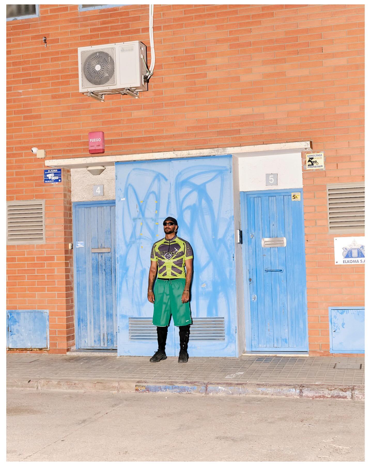 Full shot of an adult male standing in front of blue doors with graffiti on a brick building wearing casual summer clothing.