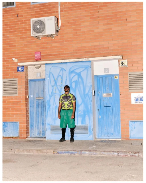 Full shot of an adult male standing in front of blue doors with graffiti on a brick building wearing casual summer clothing.