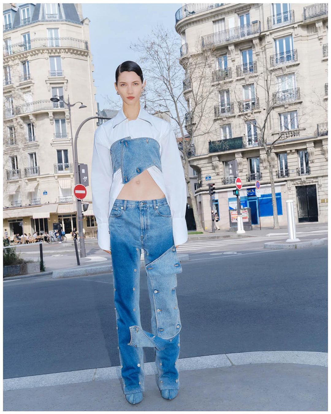 A woman posing in avant-garde denim fashion with a white blouse in an urban setting featuring Parisian buildings and city streets.