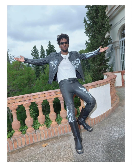 Adult male poses in leather jacket and pants on a brick patio with trees and a white building in background under an overcast sky.