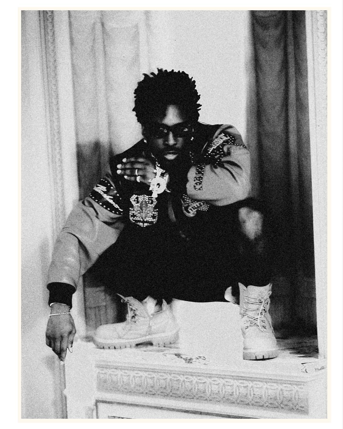 Monochrome portrait of adult male squatting indoors, wearing an ornate jacket, dark pants, work boots, and jewelry.