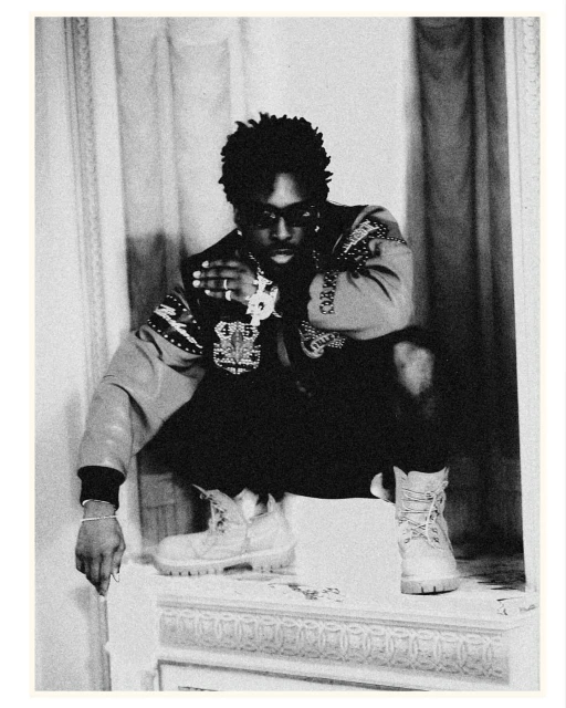 Monochrome portrait of adult male squatting indoors, wearing an ornate jacket, dark pants, work boots, and jewelry.