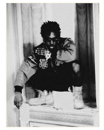 Monochrome portrait of adult male squatting indoors, wearing an ornate jacket, dark pants, work boots, and jewelry.