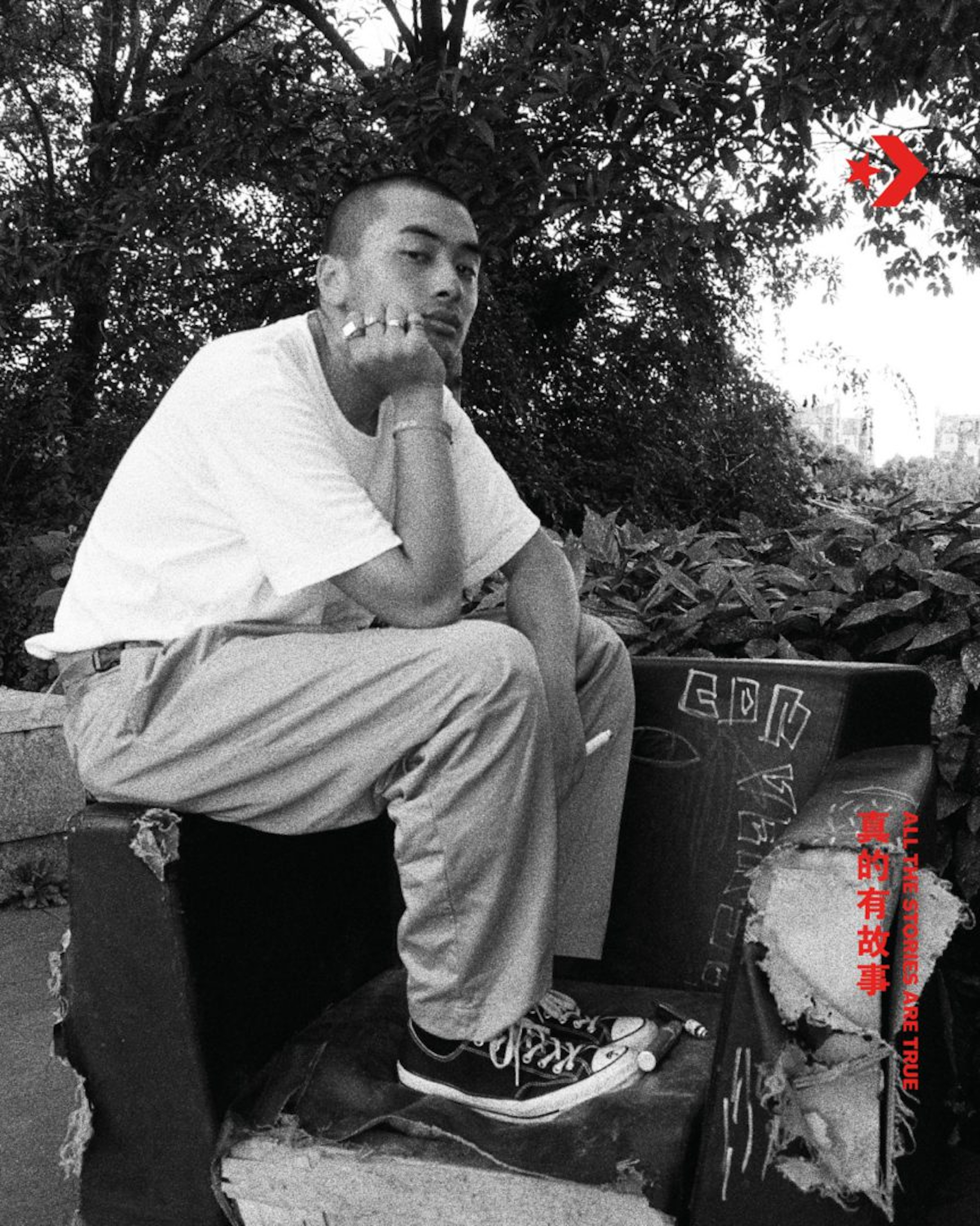 Black-and-white portrait of a young adult sitting outside on a graffiti-covered chair, Converse sneakers, and trees in the background.