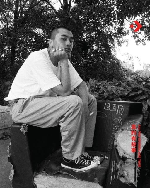 Black-and-white portrait of a young adult sitting outside on a graffiti-covered chair, Converse sneakers, and trees in the background.