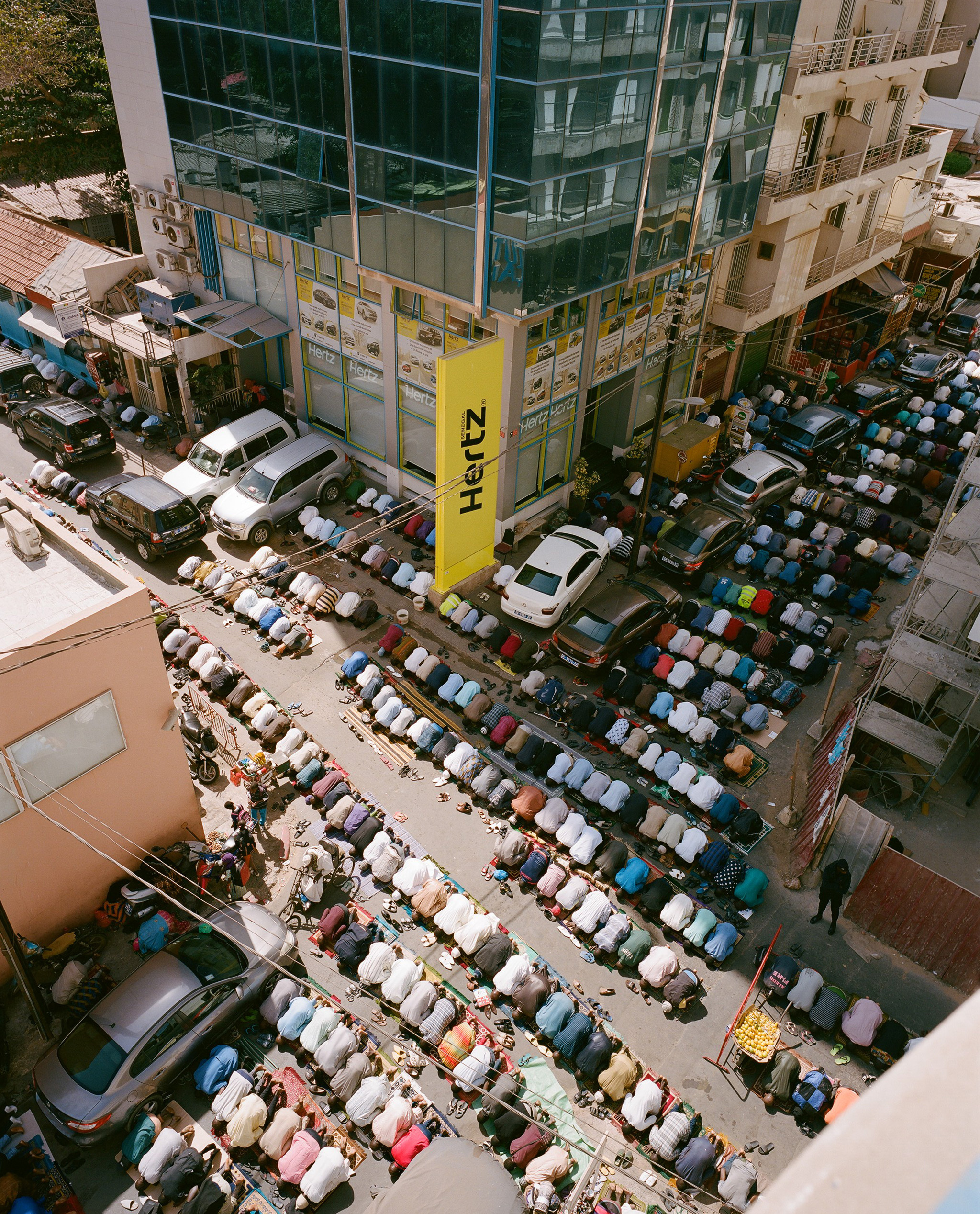Aerial view of group of adult males kneeling in prayer on street alongside parked cars and buildings under daylight conditions.