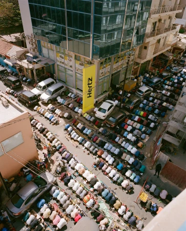 Aerial view of group of adult males kneeling in prayer on street alongside parked cars and buildings under daylight conditions.