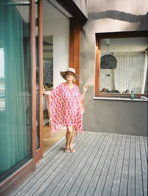 Elderly woman in patterned kaftan and sun hat stands on a wood porch in front of sliding glass doors, with sunlight and a window.