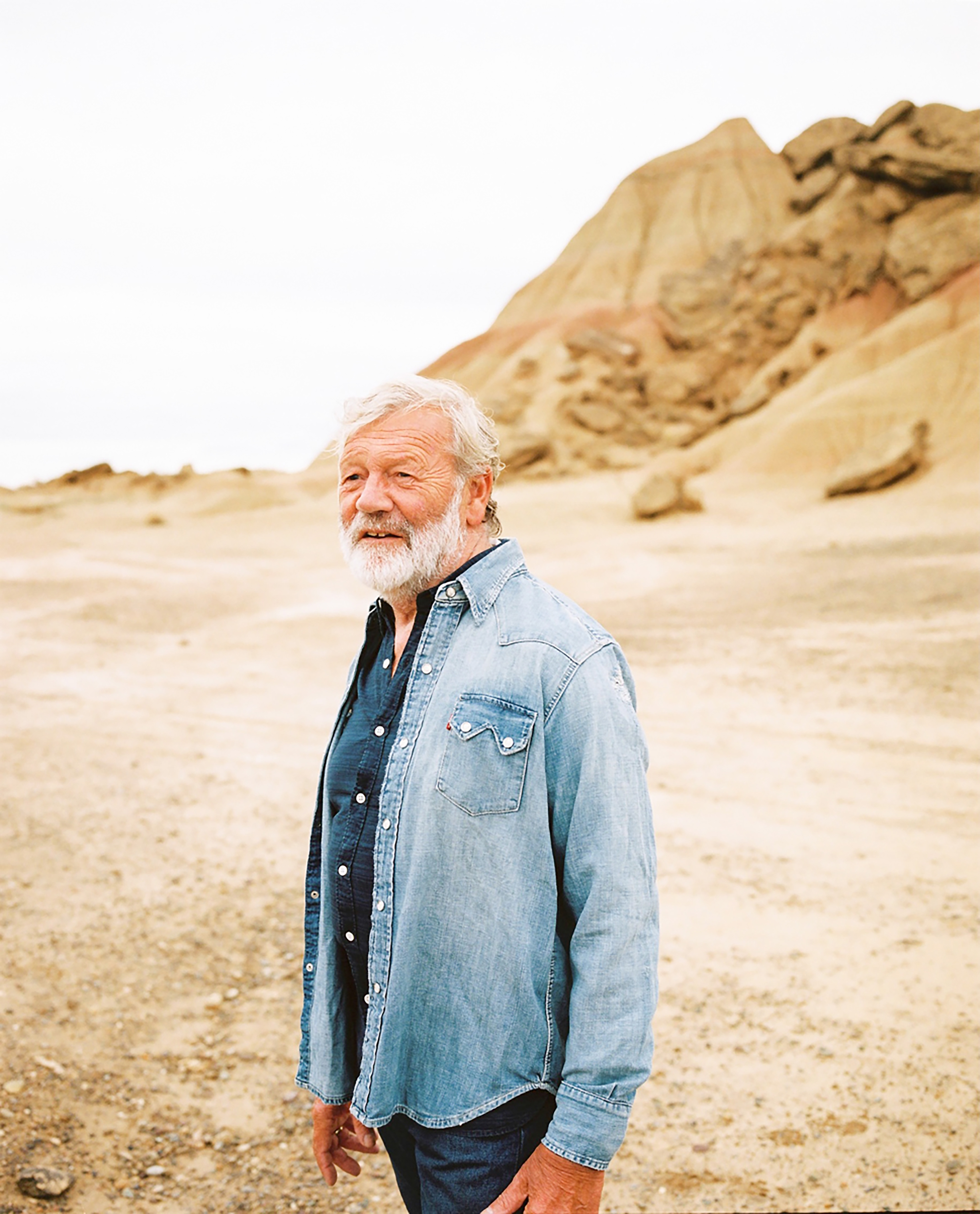 Portrait of a mature man in a denim jacket posing in a desert landscape with arid hills and a bright, overcast sky in the background.