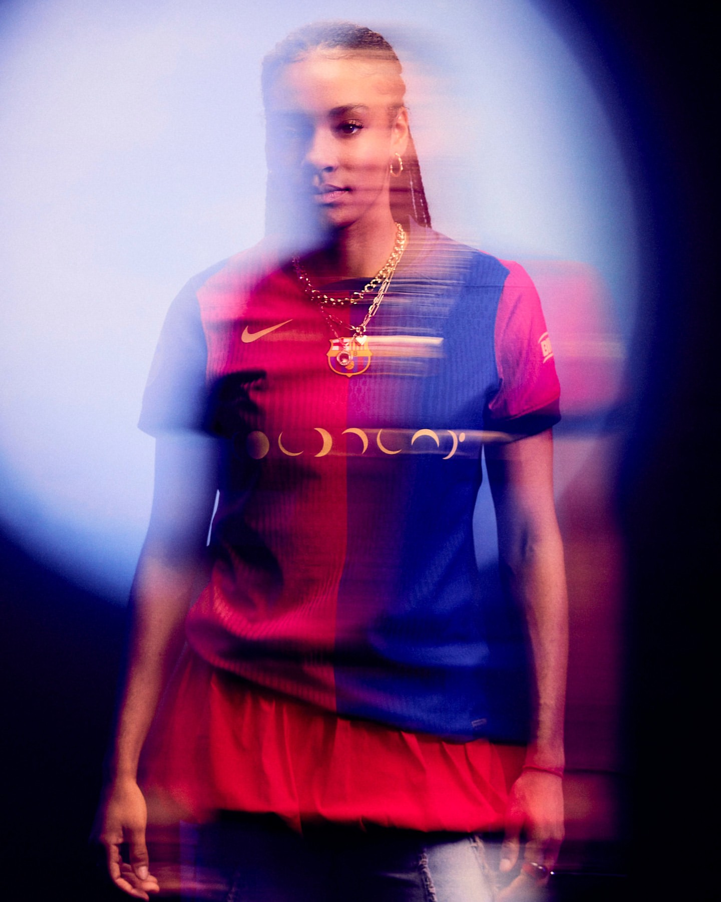 Woman in Barcelona soccer jersey, gold chain, and jeans in a blurred motion studio portrait with blue and black gradient backdrop.