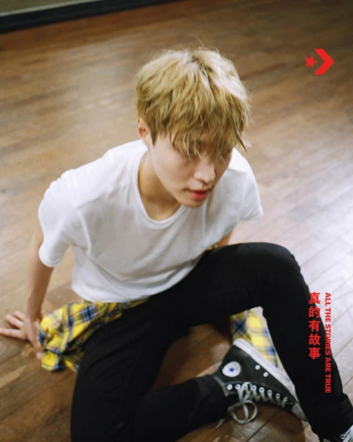 Young adult male seated on a wooden floor wearing a white t-shirt, black pants, and black and white sneakers with a Converse logo visible.