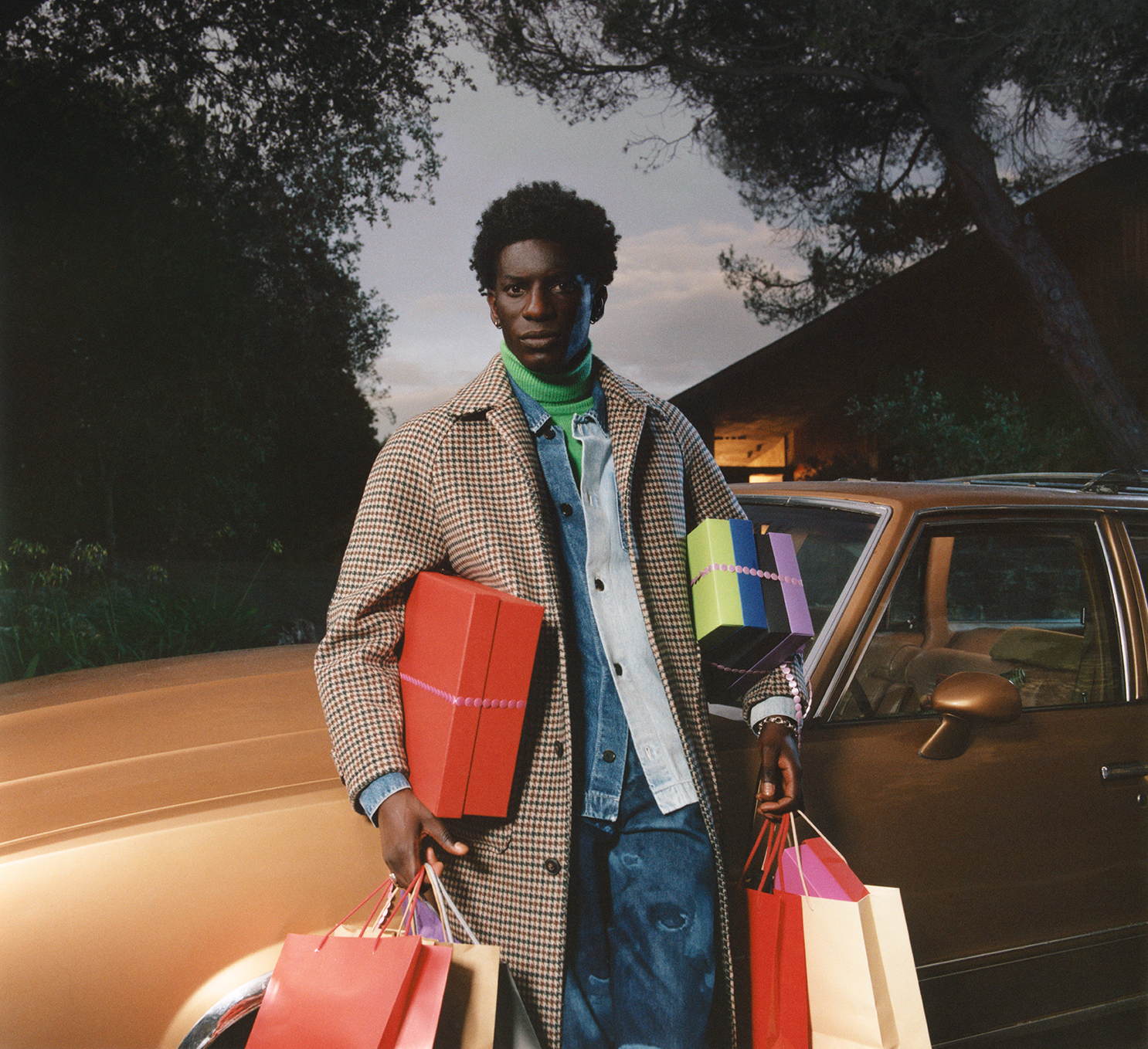 A stylish Black man outdoors stands by a vintage car, holding gifts and shopping bags, dressed in a coat, jeans, and a green turtleneck.