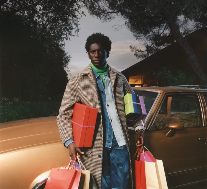A stylish Black man outdoors stands by a vintage car, holding gifts and shopping bags, dressed in a coat, jeans, and a green turtleneck.