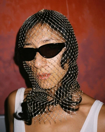 Model wearing a glittering mesh headpiece and dark sunglasses against a textured red wall background.