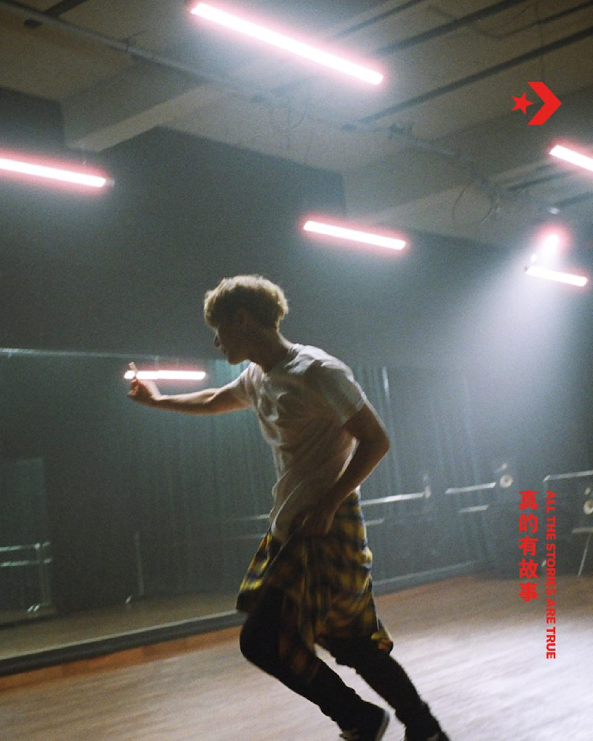 Young adult man with dyed blonde hair and a plaid shirt runs in a dance studio under bright lighting and next to a mirror.
