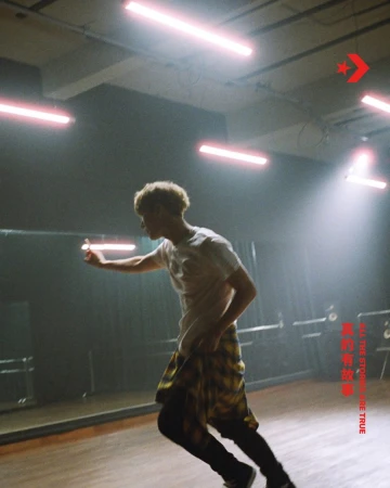 Young adult man with dyed blonde hair and a plaid shirt runs in a dance studio under bright lighting and next to a mirror.