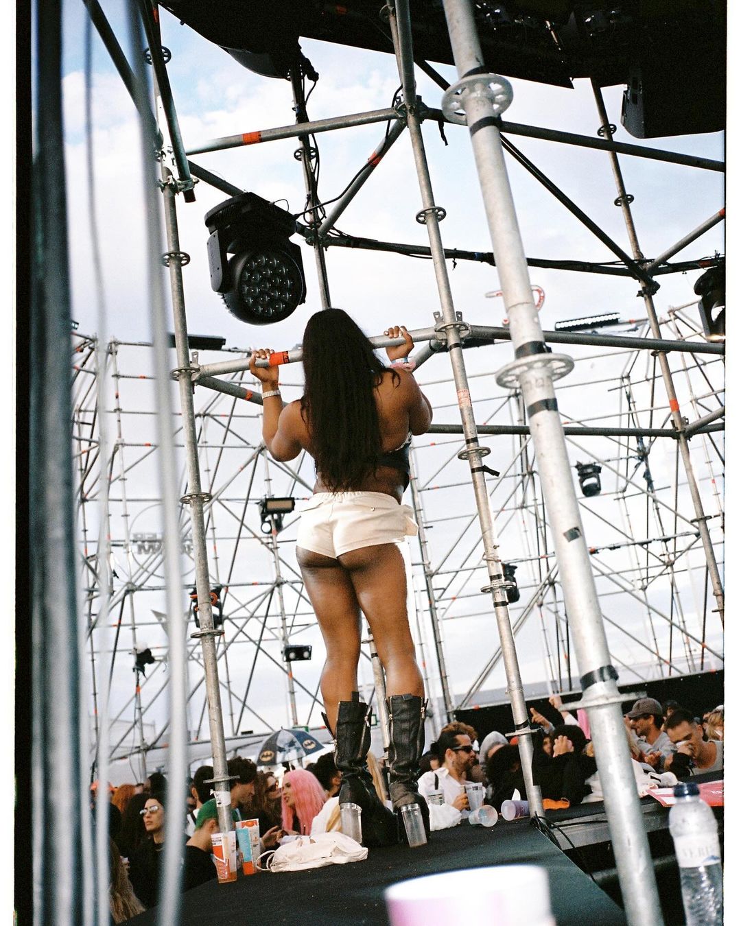 Back view of a woman on stage at a festival, wearing shorts and boots, with crowd and scaffolding in the background under a blue sky.
