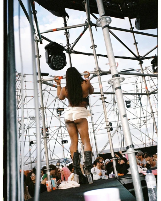 Back view of a woman on stage at a festival, wearing shorts and boots, with crowd and scaffolding in the background under a blue sky.