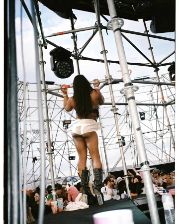 Back view of a woman on stage at a festival, wearing shorts and boots, with crowd and scaffolding in the background under a blue sky.