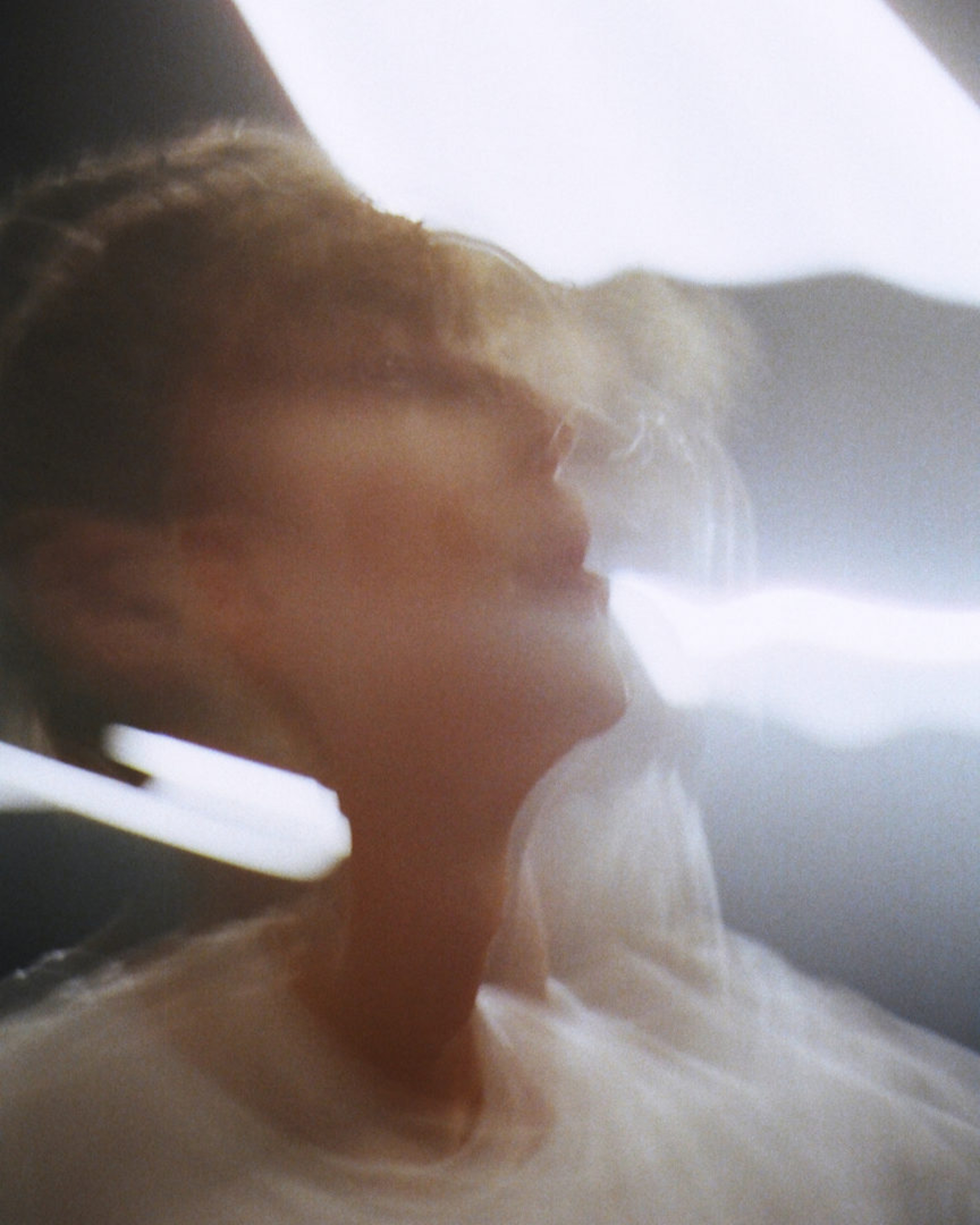 Close-up view of a person with blurred motion and neutral expression, wearing a white shirt against a bright, diffused background.