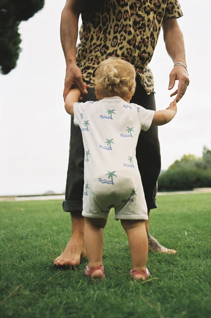 A toddler wearing a palm tree romper holds hands with an adult on a green lawn in a medium shot under an overcast sky outdoors.