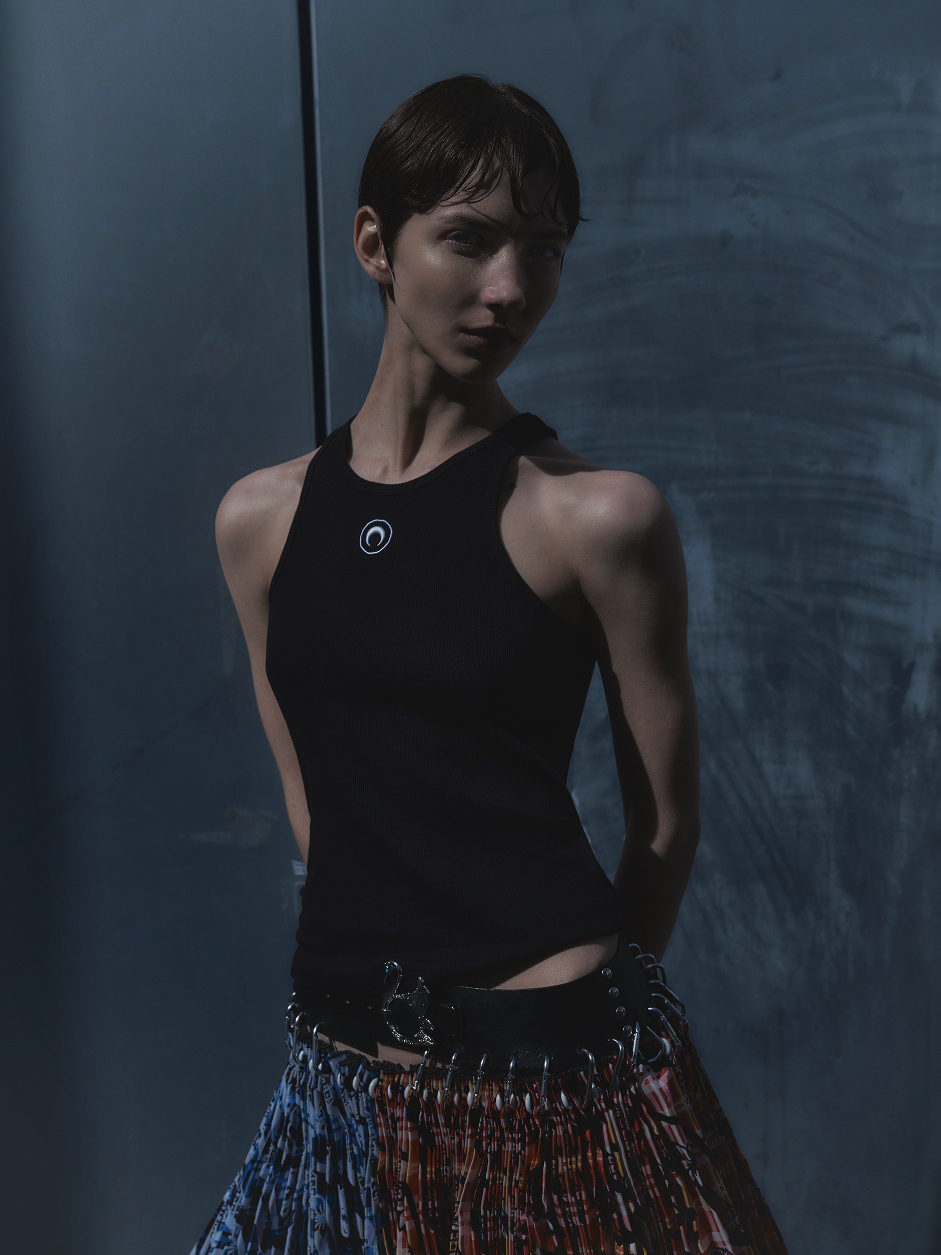 A slender woman with short dark hair poses indoors wearing a black top and colorful skirt, standing against a textured grey backdrop.