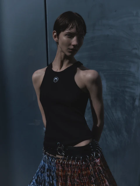 A slender woman with short dark hair poses indoors wearing a black top and colorful skirt, standing against a textured grey backdrop.
