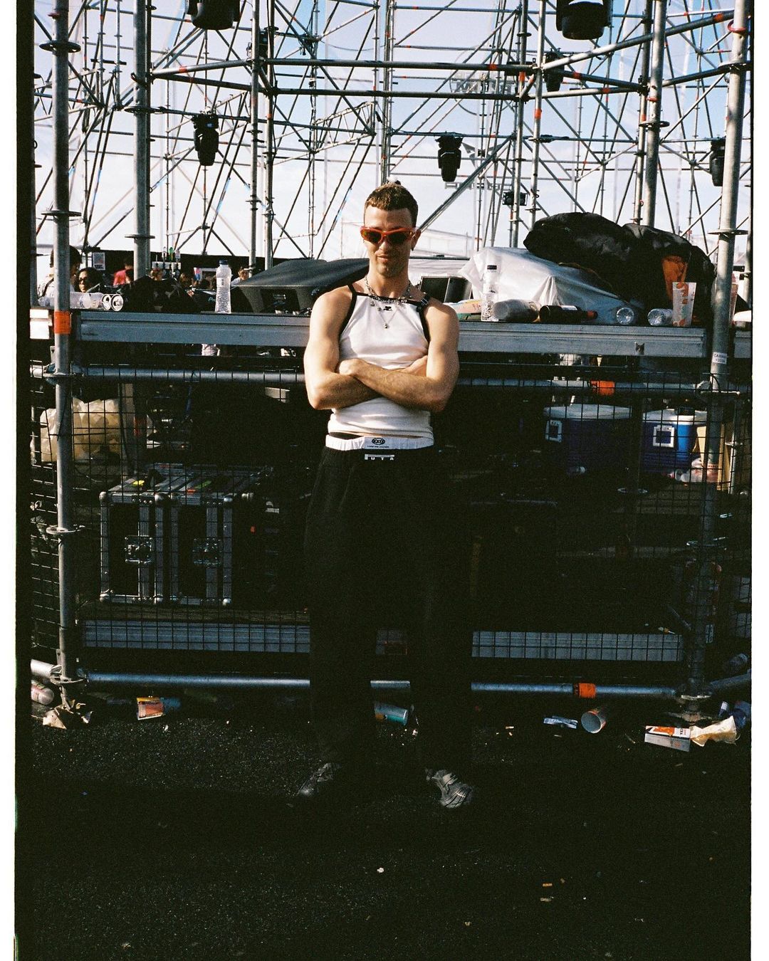 Adult male standing outdoors on an urban street in a white tank top and black pants with his arms crossed at an event stage.