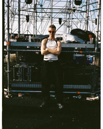 Adult male standing outdoors on an urban street in a white tank top and black pants with his arms crossed at an event stage.