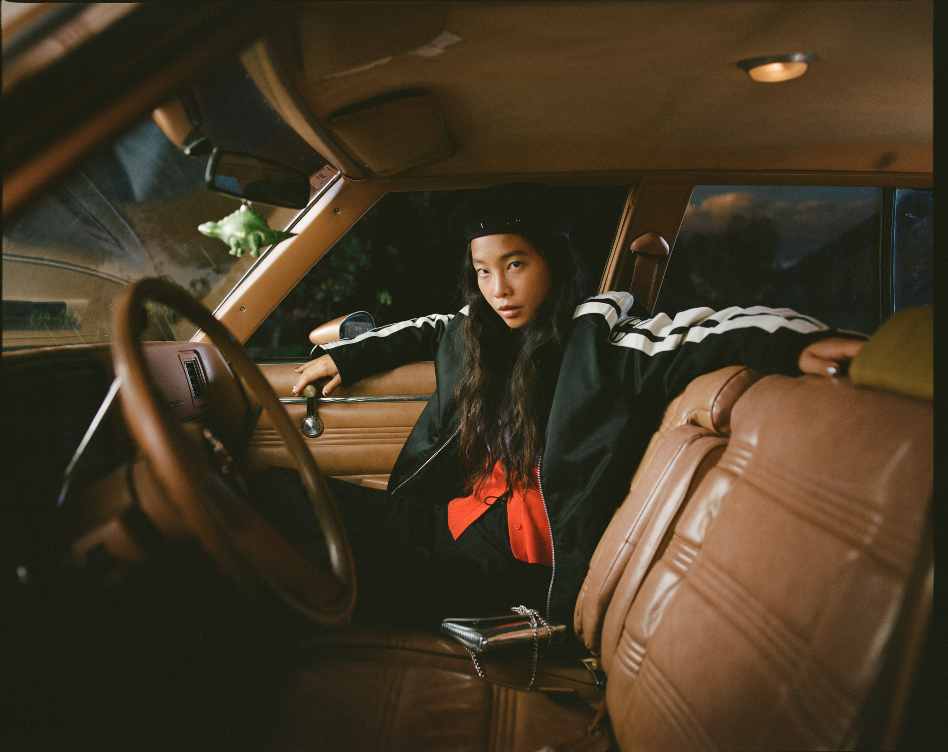 Woman in a black jacket poses inside a vintage brown car with tan leather seats, casting a moody vibe with a dinosaur toy on the mirror.