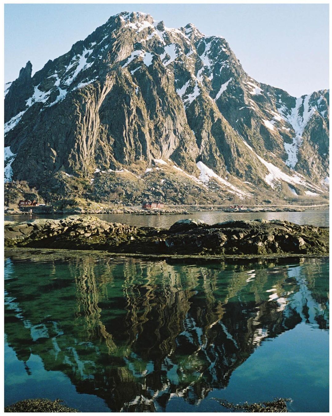Snow-capped mountain reflected in a body of water. Green islands in the foreground, with village structures at the base of the mountain.