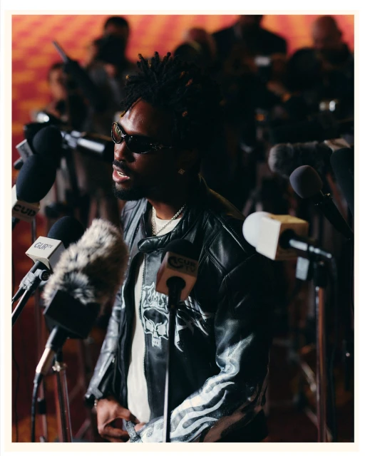 Adult male with dreadlocks wearing a black leather jacket speaks at a press conference with multiple microphones in front of him.